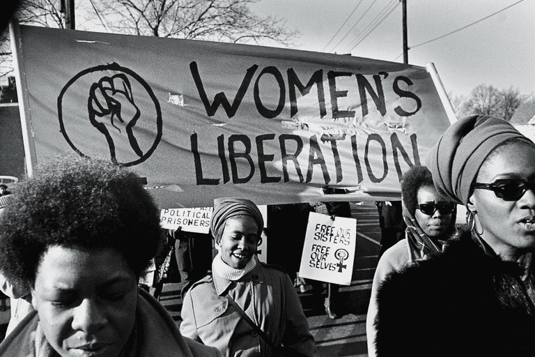 Women walk before a large banner that reads 'Women's Liberation'. Another visible, smaller sign reads: 'Free Our Sisters, Free Our Selves'