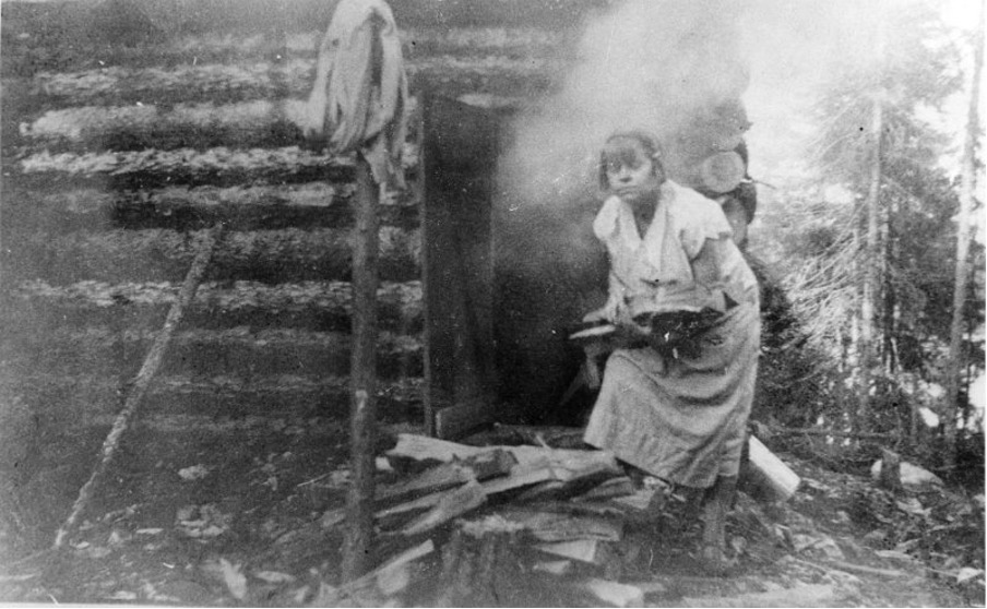 Black and white photograph of a woman loading wood into a sauna. She wears a simple dress. There are pine trees in the background.