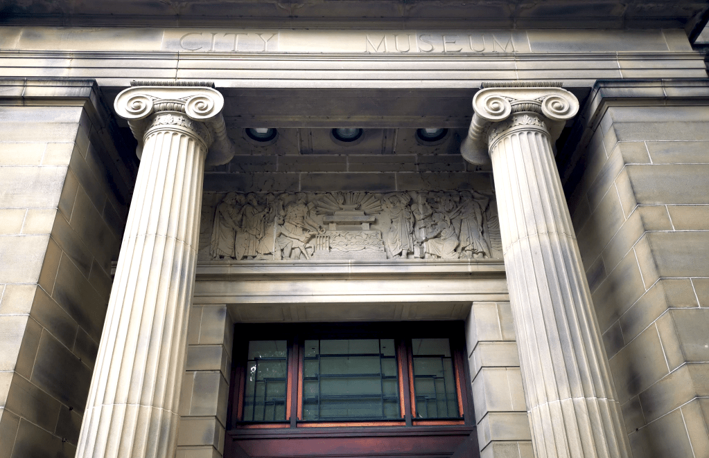 Carved frieze discussed in text. Above the door of the Weston Park Museum. Light stone, flanked by two columns and above a dark, heavy wooden door.