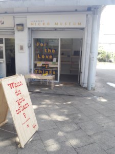 The Lansbury Micro Museum. A small museum is set up behind a shopfront. A billboard welcomes visitors in the foreground. A sign in the window reads "For the Love of Things"